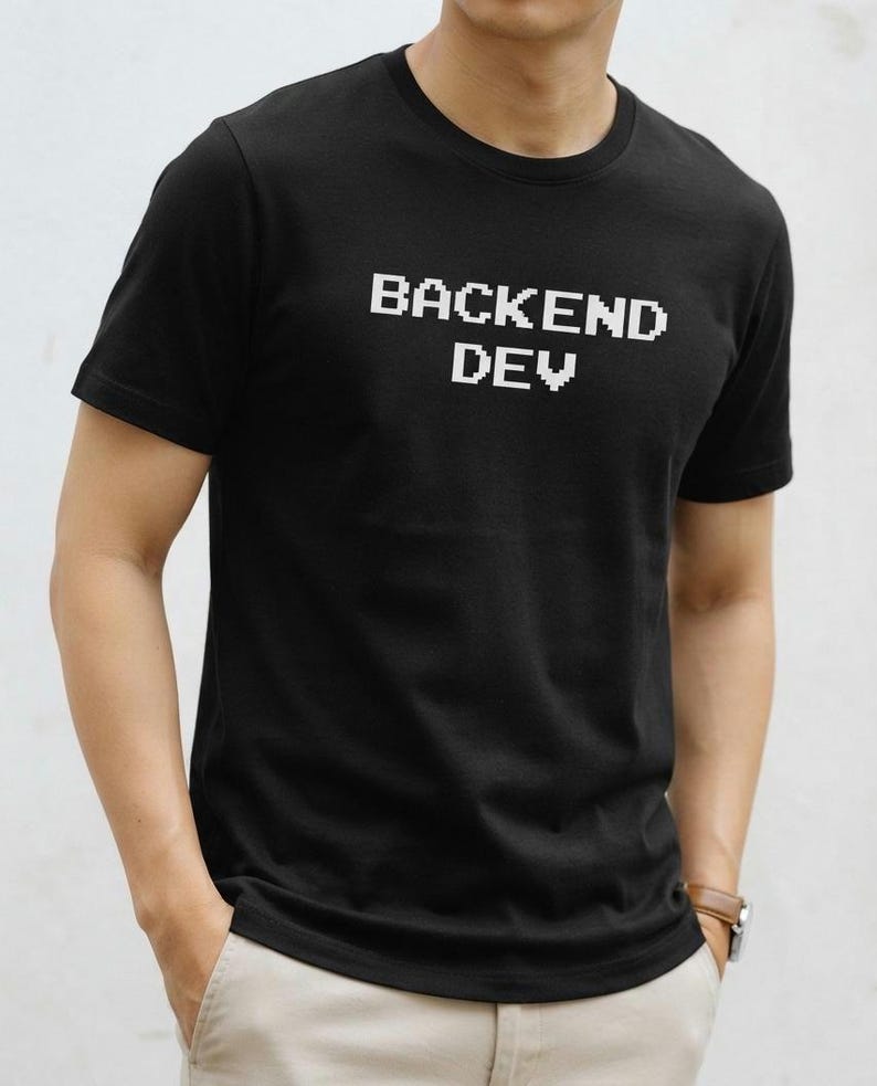 May include: A black short-sleeved t-shirt featuring the white pixelated text "BACKEND DEV" printed across the chest. The garment has a classic crew neckline and a straight hem, providing a simple and clean look for everyday casual wear.