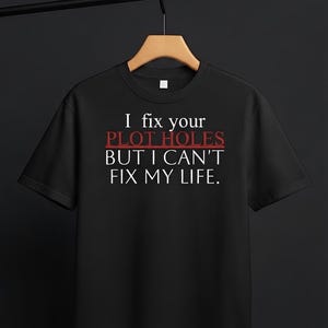 May include: A black short-sleeved t-shirt featuring white and red text that reads: I fix your PLOT HOLES BUT I CAN'T FIX MY LIFE. The shirt is displayed on a wooden hanger against a dark background, showcasing a crew neckline and a standard fit.