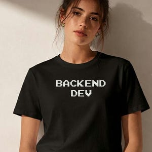 May include: A black short-sleeved t-shirt featuring the white pixelated text BACKEND DEV centered on the chest. The shirt has a classic crew neckline and is worn with blue denim jeans, showcasing a simple and functional design for everyday wear.