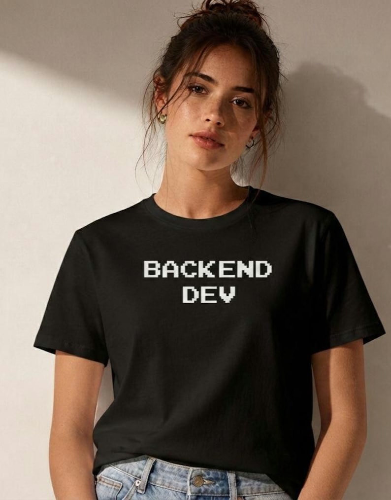 May include: A black short-sleeved t-shirt featuring the white pixelated text BACKEND DEV centered on the chest. The shirt has a classic crew neckline and is worn with blue denim jeans, showcasing a simple and functional design for everyday wear.