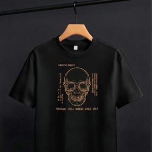 May include: A black short-sleeved t-shirt featuring a copper-coloured graphic of a skull wearing glasses. The design includes the text "SUBROUTINE ZOMBIE()" at the top and "FORTRAN: STILL UNDEAD SINCE 1957" at the bottom, with additional code-like text.