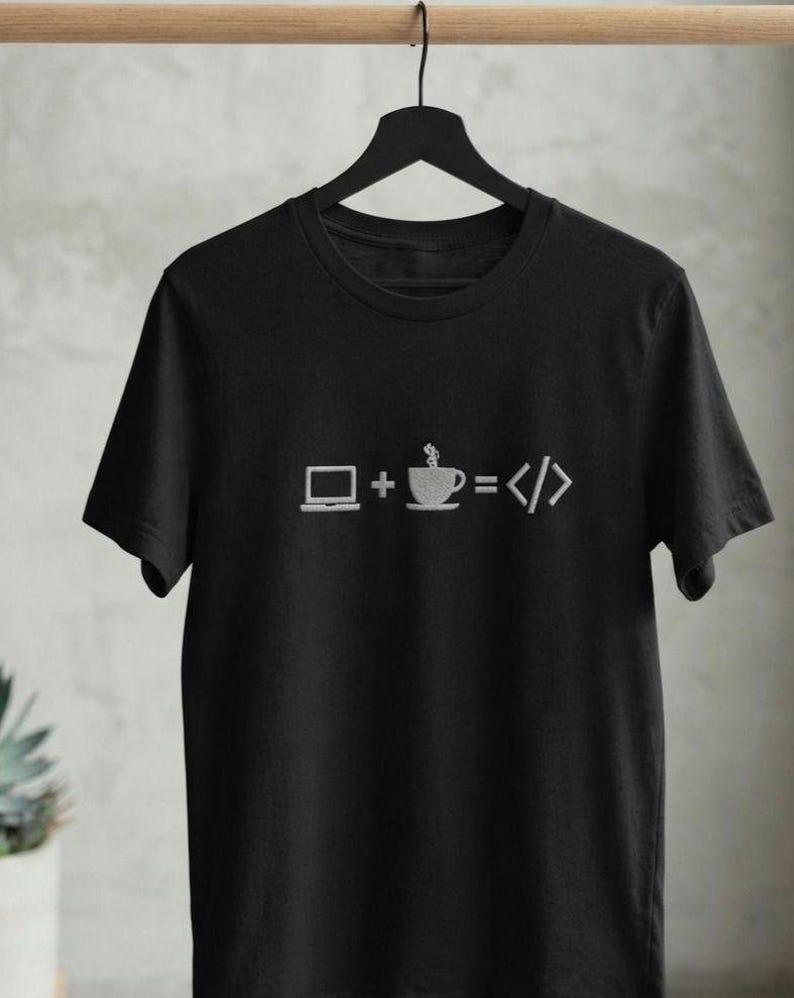 May include: A black short-sleeved t-shirt featuring a white graphic print on the chest. The design displays a laptop icon, a plus sign, a steaming coffee cup icon, an equals sign, and a code bracket symbol, representing the equation laptop plus coffee equals code.