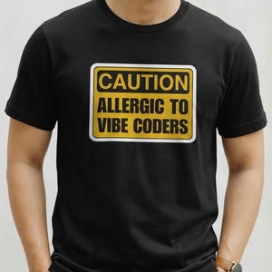 May include: A black short-sleeved t-shirt featuring a rectangular yellow sign graphic with a white border. The text on the sign is printed in bold black capital letters and reads: CAUTION ALLERGIC TO VIBE CODERS. The shirt has a classic crew neckline.