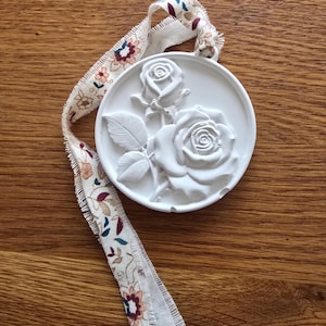 Peut inclure: Un ornement circulaire blanc présentant un relief de deux roses avec des feuilles. L'ornement est suspendu par un long ruban en tissu aux bords effilochés, décoré d'un motif floral répétitif dans des tons marron, bleu et rouge foncé.