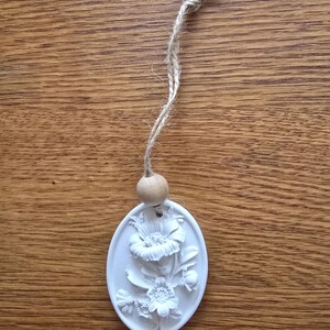 Peut inclure: Un ornement suspendu ovale blanc présentant un relief de fleurs variées. L'ornement est attaché à une ficelle de jute naturelle avec une perle en bois ronde unique positionnée directement au-dessus du sommet de la plaque florale blanche.