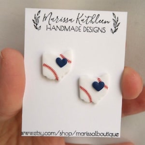 May include: Handmade earrings featuring a baseball design with a navy blue heart. The earrings are white with red stitching and are displayed on a white card with the text "Marissa Kathleen HANDMADE DESIGNS".