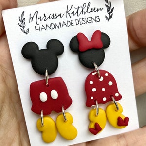 May include: A pair of dangle earrings featuring a black and red Mickey Mouse and Minnie Mouse design. The earrings are handmade and feature a red bow, white polka dots, and yellow shoes.