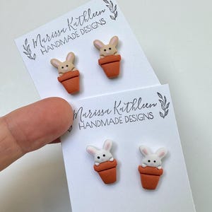 May include: Handmade bunny earrings in a terracotta pot design. The earrings are on white cards with the text "Marissa Kathleen HANDMADE DESIGNS". The earrings feature a light brown bunny with a terracotta pot.