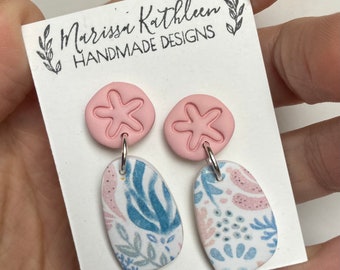 May include: A pair of pink and white dangle earrings with a starfish charm and a floral pattern. The earrings are handmade and are on a white card with the text "Marissa Kathleen Handmade Designs".