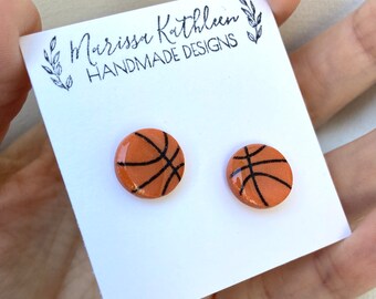 May include: A pair of round, orange basketball earrings with black line details. The earrings are displayed on a white card with the text "Marissa Kathleen Handmade Designs" in black script.