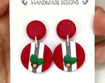 May include: A pair of red and white striped dangle earrings with a small green leaf and brown acorn charm. The earrings are handmade and have a silver hook.