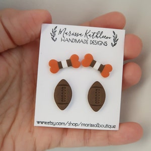 May include: A set of three pairs of earrings featuring football and bone designs. The earrings are made of polymer clay and are painted in brown, orange, and white. The earrings are displayed on a white card with the text "Marissa Kathleen Handmade Designs" and "www.etsy.com/shop/marissaboutique".