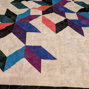 May include: A quilt featuring a repeating star pattern in shades of blue, purple, teal, and coral on a cream-colored background. The fabric displays intricate, swirling machine-stitched quilting patterns and a solid black border along the edge.