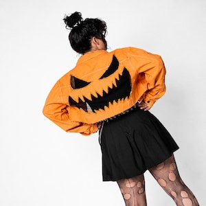 Puede incluir: Una chaqueta vaquera corta de color naranja con un gran recorte de tela negra en la espalda con forma de cara de calabaza dentada. La chaqueta tiene mangas largas, cuello y cierre de botones, combinada con una falda negra plisada.