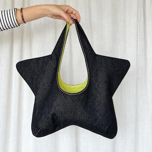 May include: A star-shaped tote bag crafted from dark blue denim fabric. The interior lining is a bright lime green colour. The bag features a single integrated handle at the top and is constructed with visible white stitching along the edges.