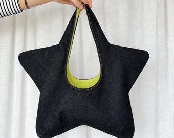 May include: A star-shaped tote bag crafted from dark blue denim fabric. The interior lining is a bright lime green color. The bag features a single integrated handle at the top and is constructed with visible white stitching along the edges.