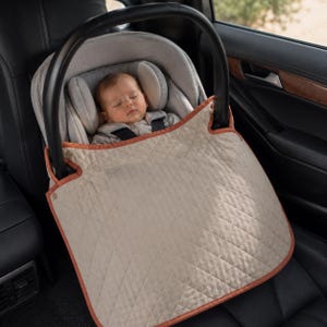 May include: A beige quilted car seat cover with a rust-colored trim attached to a black infant car seat. The cover features a diamond-stitched pattern and is secured to the handle of the seat, partially shielding a sleeping infant inside.