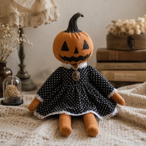 May include: A fabric doll with a round orange pumpkin head featuring a black stitched jack-o'-lantern face. The doll wears a black dress with white polka dots, white lace trim at the collar and sleeves, and a small oval pendant at the neckline.