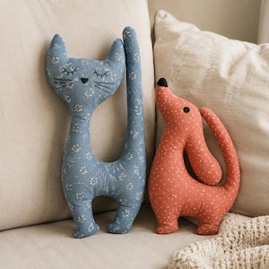 May include: A blue fabric cat plush with a white floral pattern and embroidered black facial features, standing next to a rust-coloured fabric dog plush with a white dotted pattern and a black nose, both featuring elongated, stylised shapes.