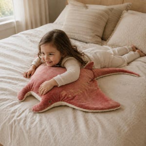 May include: A plush stingray-shaped cushion in a dusty rose colour with a cream-coloured underside. The top surface features a delicate embroidered floral pattern in a light gold thread, with a soft, velvety texture and a long, slender tail.