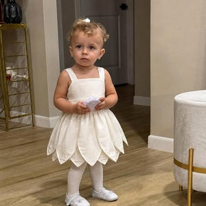 May include: A cream-coloured sleeveless dress featuring a bodice with wide straps and a skirt constructed from layered, petal-shaped fabric panels. The dress is paired with opaque white tights and white flat shoes with a small decorative bow detail.