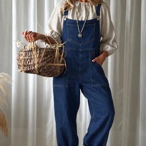 Puede incluir: Peto de mezclilla azul oscuro con bolsillo en el peto, bolsillos laterales y dobladillos vueltos. El peto se lleva sobre una blusa de manga larga a cuadros beige y blancos. Se sostiene una cesta de mimbre marrón con trigo seco y flores.