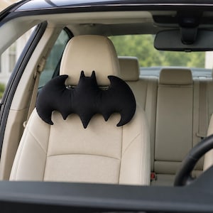 May include: A black, plush, bat-shaped headrest pillow attached to the beige headrest of a car seat. The pillow features the iconic pointed ears and scalloped wing design of a bat, providing a soft, cushioned support for the neck area.