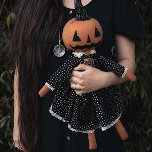 May include: A fabric doll featuring an orange pumpkin head with a black painted jack-o'-lantern face and a black stem. The doll wears a black dress with white polka dots, white lace trim at the collar and cuffs, and a small round gold-toned brooch.
