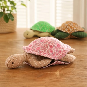 Pode incluir: Três tartarugas de tecido acolchoadas. A tartaruga em primeiro plano tem uma carapaça com padrão floral rosa e um corpo castanho-claro com riscas brancas. Ao fundo, uma tartaruga tem uma carapaça floral verde e outra uma carapaça castanha.