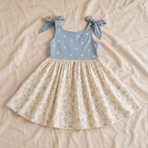May include: A sleeveless dress featuring a dusty blue bodice with a small white daisy print and tie-shoulder straps. The skirt is a cream-colored fabric adorned with a dense pattern of tiny blue and green floral sprigs, creating a flared silhouette.