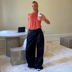 May include: A coral-colored sleeveless halter top with a high neckline and a gathered bodice paired with dark navy blue wide-leg trousers featuring a pleated front and a high-waisted fit. The outfit is completed with light-colored athletic sneakers.