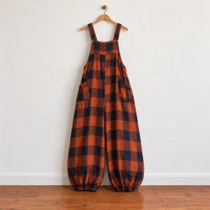 May include: A pair of overalls featuring a large-scale orange and navy blue buffalo plaid pattern. The garment has wide shoulder straps, a square neckline, two side pockets, and loose-fitting legs that taper into gathered elasticized cuffs.