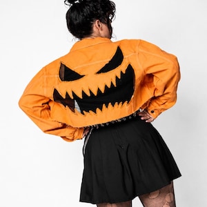 May include: An orange cropped denim jacket featuring a large, jagged-edged black fabric cutout on the back that forms a stylized jack-o'-lantern face with sharp, triangular eyes and a wide, toothy grin. The jacket has a raw, frayed hemline.