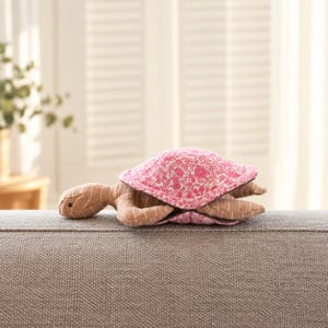 Puede incluir: Un juguete de peluche con forma de tortuga que presenta cabeza y extremidades de tela color canela con rayas blancas, junto a un caparazón rosa vibrante con pequeños motivos florales en blanco y rosa oscuro. La tortuga está en posición horizontal.