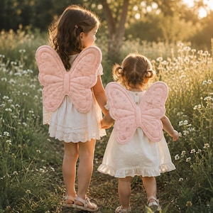 May include: Two pairs of soft, quilted light pink butterfly wings with a subtle floral pattern, worn by two individuals dressed in white dresses. The wings feature a distinct butterfly shape with stitched detailing and are secured on the back.