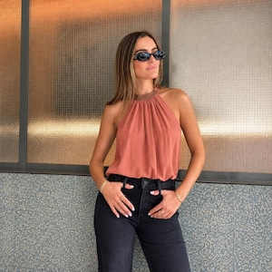Può includere: Un top all'americana senza maniche color terracotta con scollo alto e fascia del colletto in bronzo metallizzato, abbinato a jeans neri a vita alta. Il top presenta un tessuto morbido e drappeggiato, mentre i jeans hanno una classica chiusura a bottoni.