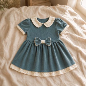 May include: A teal corduroy dress featuring short puff sleeves, a rounded collar with a cream and yellow floral pattern, and a matching floral trim on the sleeve cuffs and hem. A central teal corduroy bow sits at the waistline of the dress.