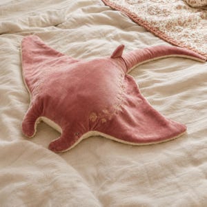 May include: A plush stingray-shaped pillow in a dusty rose velvet fabric. The top surface features delicate, light-colored floral embroidery along the center and wings. The underside and edges are lined with a soft, cream-colored textured material.