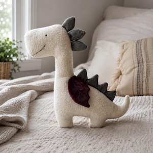 May include: A plush dinosaur toy made of cream-colored textured fabric. It features a dark maroon velvet wing, black and white patterned fabric spikes along its back and head, and a simple stitched mouth and eye detail on its side profile.