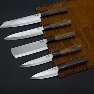Pode incluir: Um conjunto de cinco facas de cozinha com lâminas de metal prateado e cabos de madeira castanho-escuro. Cada lâmina apresenta um reforço preto e os caracteres kanji japoneses para Osaka gravados perto da base das facas alinhadas.