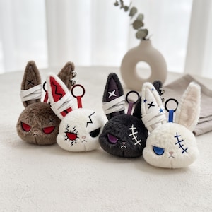 May include: Four plush rabbit keychains featuring faux fur, embroidered facial expressions, and stitched details. Each rabbit has one ear wrapped in white fabric strips and a circular metal ring attached to the top for hanging purposes.