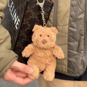 May include: A light brown, fuzzy plush teddy bear keyring featuring a silver-toned metal clasp and chain. The bear has a soft, textured surface, small rounded ears, and a darker brown stitched nose, attached to a dark brown patterned bag.