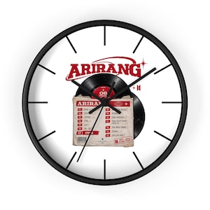 Puede incluir: Un reloj de pared redondo con marco negro y esfera blanca que presenta un gráfico de un disco de vinilo negro y una tarjeta de lista de canciones etiquetada como ARIRANG. El reloj tiene manecillas negras. Incluye texto y detalles rojos.