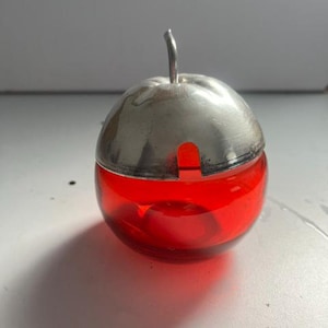 Op de afbeelding: Een ronde, doorschijnende rode glazen houder in de vorm van een appel, voorzien van een afneembaar zilverkleurig metalen deksel met een gebogen steeltje als handvat en een kleine inkeping aan de zijkant voor een lepel.
