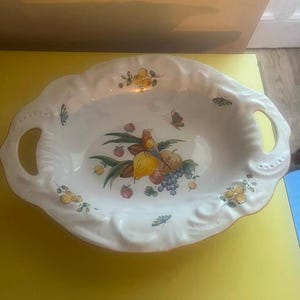 Peut inclure: Un bol de service ovale en céramique blanche avec deux poignées intégrées et un bord festonné. L'intérieur présente une illustration peinte colorée d'un oiseau perché sur une poire, entouré de raisins, de baies et de petits papillons.