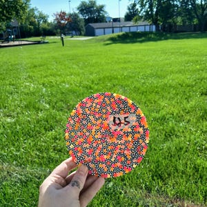 May include: A circular coaster crafted from small, fused plastic beads in a vibrant pattern of neon orange, pink, and yellow dots interspersed with black and white star-shaped accents. A small white sticker with the handwritten text $25 is attached.
