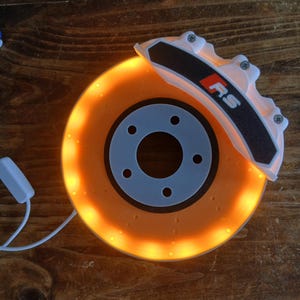 Op de afbeelding: Een ronde lamp in de vorm van een remschijf voor auto's met een witte remklauw waarop de tekst RS in rood en wit staat. De schijf geeft een warm oranje licht af langs de rand en is voorzien van een wit stroomsnoer met een kleine schakelaar.