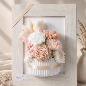 Peut inclure: Un cadre photo en bois clair contenant un arrangement floral en trois dimensions dans des tons blanc, crème et pêche doux. Les fleurs sont fixées dans une base blanche semi-circulaire côtelée avec un ruban beige. Une étiquette indique TRIO MAR atelier.