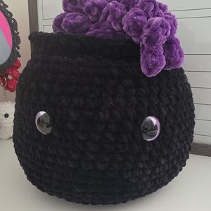 May include: A round, black crocheted cauldron with a textured, plush surface. It features two large, glossy black eyes with purple rims and is filled with a cluster of deep purple, soft, curly yarn that spills over the top edge of the container.