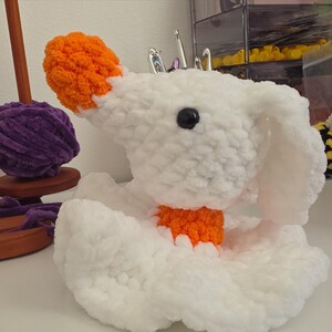 May include: A white crocheted plush toy featuring a rounded head with a bright orange nose, a black plastic eye, and long floppy ears. The figure has a small orange collar around its neck and a ruffled white base, crafted from soft yarn.